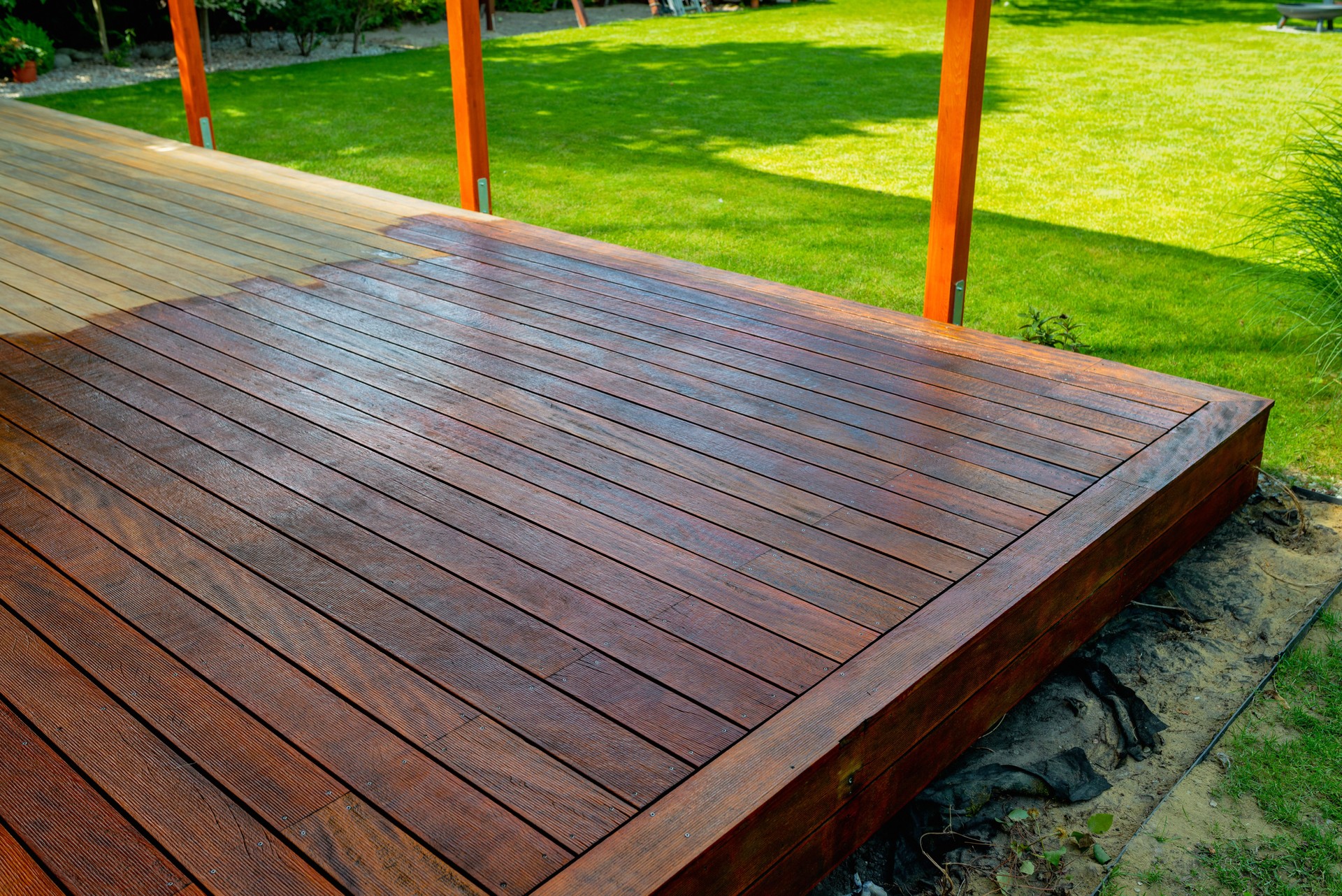 Close-up of a wooden deck showing the process of restoration with a half-stained surface contrasting weathered and treated wood during a home improvement project - in the background a well-kept garden